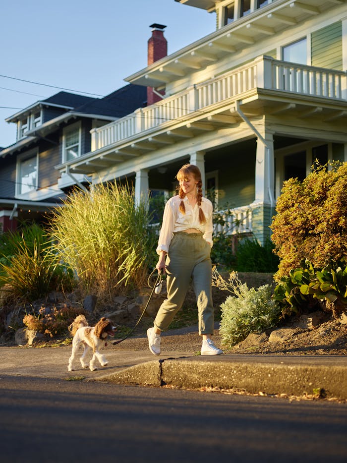 A smiling woman walks her dog in a sunlit suburban neighborhood, enjoying leisure time.